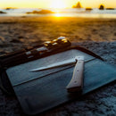The Messermeister Adventure Chef Maple Folding Flexible Fillet Knife - 6" with a metallic handle rests on its closed case atop a rock at the beach, crafted from German stainless steel, as the sun sets over ocean rocks in the background.
