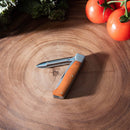 The Messermeister Adventure Chef Maple Folding Peeler & Parer, featuring a wooden handle and partially serrated blade, is open on a wooden cutting board with tomatoes and leafy greens in the background.