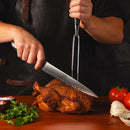 Wearing a dark apron, a person uses the Messermeister Avanta Damascus knife and fork to carve roasted chicken on a wooden table, surrounded by parsley, tomatoes, garlic, and salt.