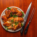 A plate of roasted, sliced chicken garnished with parsley rests on a wooden table beside a Messermeister Avanta Damascus Block Set carving knife and meat fork.