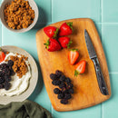 A wooden cutting board with strawberries, blackberries, and the Messermeister Avanta Damascus Starter Set - 3 Piece chef’s knife sits on blue tiles. Nearby are bowls of granola and yogurt topped with berries.