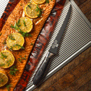 A cooked salmon fillet with lemon and dill sits on a baking sheet beside a Messermeister Avanta Kendrick BBQ Fillet Knife - 7", atop a wooden surface.