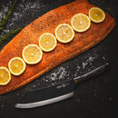 A seasoned raw salmon fillet topped with six lemon slices, coarse salt, and rosemary sits on a dark surface beside the Messermeister Avanta Kendrick BBQ Knife - 8" by Messermeister.