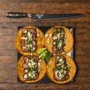 Four steak tacos with crumbled cheese and herbs are served on a black plate with lime wedges. Above, a Messermeister Avanta Kendrick BBQ Scalloped Brisket Knife – 10" rests on a rustic wooden table.