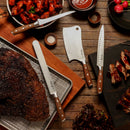 A top-down view of a wooden table displays the Messermeister Avanta Pakkawood Flexible Boning Knife (7"), along with other kitchen knives, a cutting board, cooked brisket, ribs, chicken wings, dipping sauces, spices, and salad.