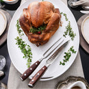 A roasted turkey with fresh herbs rests on a white platter beside the Messermeister Avanta Pakkawood Kullens Carving Set - 2 Piece. Plates, utensils, and glasses are set, ready for a memorable meal.