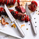 The Messermeister Avanta Stainless Fine Edge Steak Knife Set (4 Piece) features sleek stainless steel handles, displayed on a white plate beside skewered grilled meat and vegetables, with scattered peppercorns highlighting the scene.