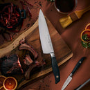 The Messermeister Blacksmith Chef's Knife - 8", crafted from German stainless steel, lies on a cutting board with cooked ribs, peppercorns, dried chilies, orange slices, a paintbrush, and parsley—all artfully arranged on a brown surface.