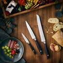 A wooden table displays slices of bread with three Messermeister Blacksmith Scalloped Slicing Knives (8.5"). Above, a wooden tray holds cheese, fruit, charcuterie; below, a green plate has salami, prosciutto, cheese, grapes, and berries. Leaves decorate the scene.