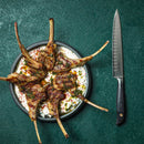 A plate of grilled lamb chops on creamy yogurt sauce, garnished with herbs and spices, sits beside a Messermeister Blacksmith Kullens Carving Knife - 9" crafted from German stainless steel.