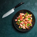 A black bowl of watermelon, grilled halloumi, and basil sits on a dark surface next to the Messermeister Blacksmith Kullens Santoku Knife - 6.5", expertly crafted from German stainless steel.