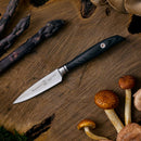 The Messermeister Blacksmith Paring Knife - 3.5" with a black handle rests on wood, surrounded by purple asparagus and mushrooms.