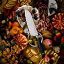 The Messermeister Blacksmith Kullens Santoku Knife (6.5") by Messermeister lies on a platter filled with crackers, olives, cheeses, meats, berries, sliced fruits, pastries, and flowers for a vibrant charcuterie spread.
