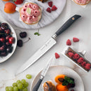 The Messermeister Blacksmith Scalloped Slicing Knife - 8.5" is displayed on a table with a paring knife, cakes, raspberries, grapes, cherries, an apricot, and a chocolate pastry topped with raspberries and mint.