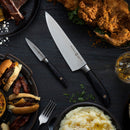 A chef’s knife and the Messermeister Blacksmith Paring Knife - 3.5" rest on a dark table, surrounded by fried chicken, mashed potatoes with pepper, burgers, a fork, and a glass of whiskey.