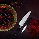 A berry tart rests on a dark table beside the Messermeister Blacksmith Chef’s Knife - 8". Nearby, red rose petals, a rose, strawberries, and cherries create an elegant scene.