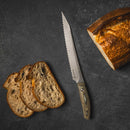 The Messermeister Carbon Scalloped Knife - 9" rests beside three slices of rustic bread and a partially sliced loaf on a dark, textured surface.