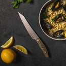 A bowl of pasta with mussels sits beside the Messermeister Carbon Utility - 6" kitchen knife on a dark surface, accompanied by a whole lemon, two lemon wedges, and scattered parsley leaves.