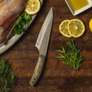 The Messermeister Chef April Bloomfield 6.5" Carbon Chef's Knife rests on wood, surrounded by lemon slices, fresh herbs, a parsley-topped fish platter, and a dish of olive oil.