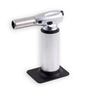 The Messermeister Cheflamme Culinary Torch by Messermeister features a silver metal body, black ignition knob, and textured grip, standing upright on a flat black base against a white background.