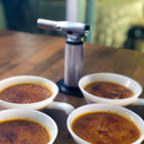 Four ramekins of crème brûlée with caramelized tops sit on a wooden table, with the Messermeister Cheflamme Culinary Torch by Messermeister placed in the background.