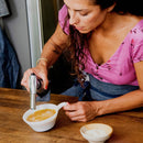 A woman in a pink shirt uses the Messermeister Cheflamme Culinary Torch by Messermeister to caramelize a dessert in a white ramekin on a wooden table, with a small bowl of sugar beside her.