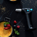 The Messermeister Cheflamme Mini Culinary Torch rests on a black surface beside crème brûlée—one topped with berries and mint arranged as a smiling face—with scattered brown sugar crystals nearby.