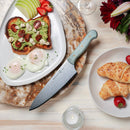 A breakfast spread of avocado toast with cherry tomatoes and feta, poached eggs, green apple, strawberries, croissants, a Messermeister Custom Blue Chef's Knife - 8", and a candle on a light wooden table.