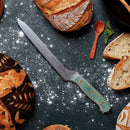 The Messermeister Custom Blue Scalloped Offset Knife - 8" rests on a dark, flour-dusted surface amid artisan bread, a wooden spoon, and mint sprig.