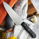 The Messermeister Custom Chef's Knife - 8" with an ergonomic handle rests on a striped towel beside a raw salmon fillet, wooden cutting board, garlic, lemon, and oil—all arranged on a stone surface.