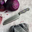 A Messermeister Custom Kullens Santoku Knife - 7" lies on a textured gray surface next to a whole red cabbage and a jar of pickled red cabbage.