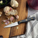 A Messermeister Custom Paring Knife - 3.5" with a black handle sits on a wooden cutting board beside garlic, with a striped towel and red onion nearby—ideal for precision prep.
