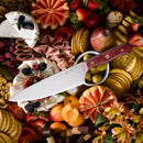An 8" Messermeister Custom Red Chef's Knife with an ergonomic handle rests on a charcuterie board adorned with assorted cheeses, crackers, fruits, olives, meats, and flowers for an artistic presentation.