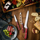 A wooden table features a cheese and charcuterie board with grapes, salami, strawberries, bread slices, scattered nuts, and three knives—including the Messermeister Custom Red Scalloped Offset Knife – 8" with its signature red handle.