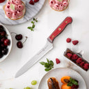 The Messermeister Custom Red Scalloped Offset Knife - 8" features a vibrant red handle and rests on a white surface, surrounded by desserts, fresh cherries, grapes, raspberries, and mint leaves.