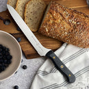 A Messermeister Custom Scalloped Offset Knife - 8" rests on a striped cloth next to sliced multigrain bread on a wooden board, with a bowl of blueberries and scattered berries nearby.