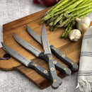 The Messermeister Custom Steak Knife Set - 4 Piece features sleek black handles and sharp steel blades, displayed on a wooden cutting board with fresh asparagus, garlic bulbs, and a striped kitchen towel on a gray countertop.