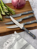The Messermeister Custom Steak Knife Set—four razor-sharp steel knives with black handles—rests diagonally on a wooden cutting board next to fresh asparagus, half a red onion, and a cream-striped cloth.