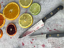 Two knives, including the Messermeister Custom Utility Knife - 6" with dark handles, rest on a gray surface among orange, lemon, and blood orange slices. Red juice stains are visible near the Messermeister utility knife and fruit.
