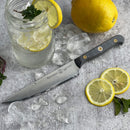 The Messermeister Custom Utility Knife - 6" by Messermeister rests on a gray surface beside a mason jar of lemon ice water, a whole lemon, two lemon slices, mint leaves, and scattered ice cubes.