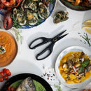 A pair of Messermeister Esther Choi Push Button Take-Apart Precision Shears - 7" sits on a white surface, surrounded by plates of seafood, gnocchi in yellow sauce, whole fish in green sauce, oysters, red soup, fresh herbs, and cherry tomatoes.