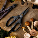 The Messermeister Esther Choi Push Button Take-Apart Precision Shears - 7" rest on a wooden board, surrounded by assorted mushrooms and purple asparagus, with moss in the corner.
