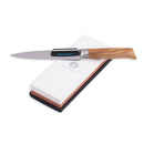 A kitchen knife with a wooden handle rests on the Messermeister Maintenance Stone, featuring 1000 & 3000 grit surfaces, a white top with the Messermeister logo, and a black base. The silver blade lies across this dual-sided sharpening stone.