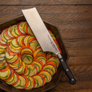 The Messermeister Meridian Elite Nakiri Knife - 6.5", featuring a black handle, lies on a pan of neatly arranged zucchini, yellow squash, and tomato slices with herbs—all displayed on a wooden table.