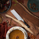 The Messermeister Meridian Elite Nakiri Knife - 6.5" is displayed on a wooden cutting board with autumn accents like pumpkin pie, whipped cream, pecans, cinnamon sticks, dried oranges, and nuts, highlighting its German-forged blade.