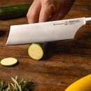 A hand slices a green zucchini with the Messermeister Meridian Elite Nakiri Knife - 6.5" on a wooden cutting board; this Messermeister essential gleams as fresh herbs are scattered nearby.