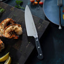 The Messermeister Meridian Elite Chef and Parer Set rests on a cutting board beside grilled shrimp halves, chopped herbs, and lemon wedges, with a blue plate and roasted tomatoes in the background.