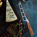 A wedge of blue cheese on a black platter with figs, grapes, crackers, and the Messermeister Oliva Elite Cheese & Tomato Knife - 5", featuring an olive wood handle, all arranged on a dark blue textured surface.