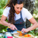 A woman in a gray apron and white shirt slices orange bell peppers outdoors using the Messermeister Oliva Elite Chef's Knife - 6" on a wooden cutting board, with a stainless steel bowl nearby.