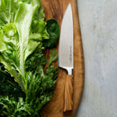 The Messermeister Oliva Elite Chef's Knife - 9" with a hand-forged blade and olive wood handle rests on a wooden cutting board beside fresh lettuce and dill, set on a light surface.